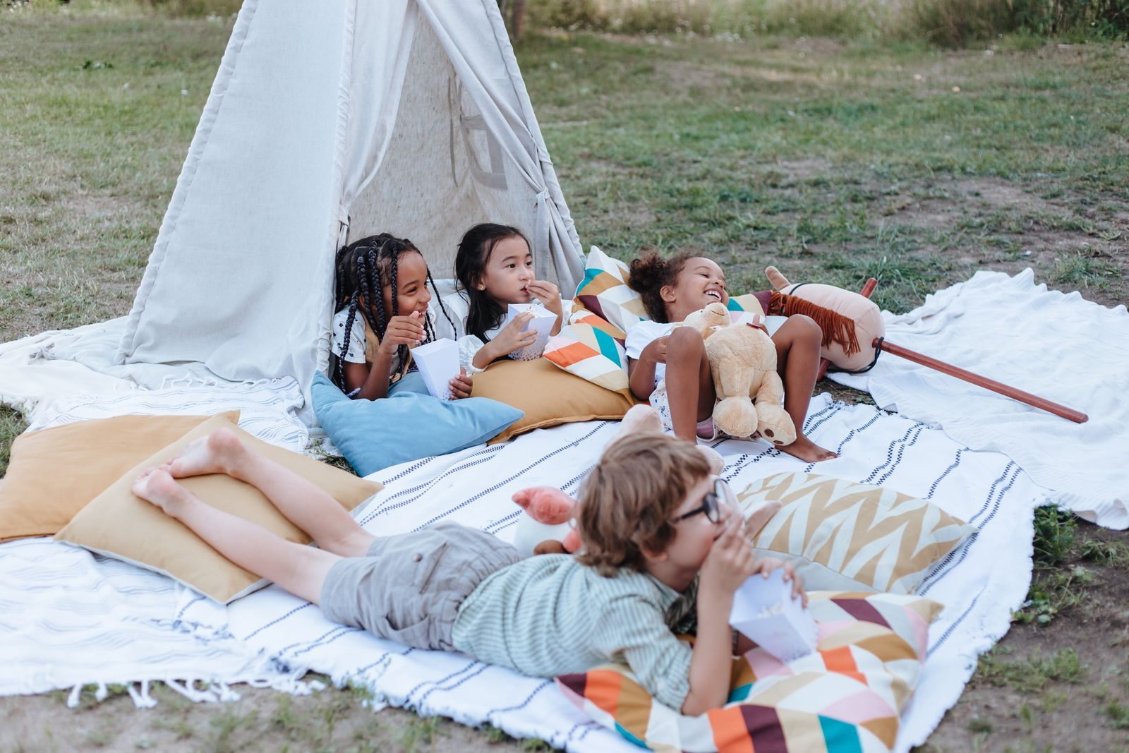 Children lying on blankets and pillows around a teepee in a backyard, a classic among staycation ideas for kids