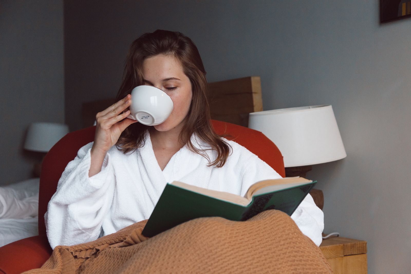 Woman in a bathrobe sipping coffee and reading a book in a cozy armchair